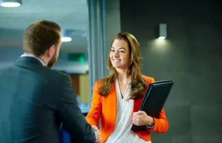 Woman in orange blazer shakes hands, holding a folder.