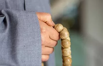Person in gray robe holding wooden prayer beads.