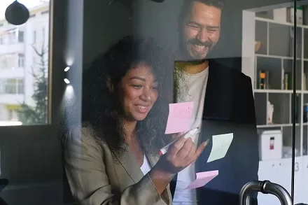 Two colleagues smiling while brainstorming with sticky notes on glass.