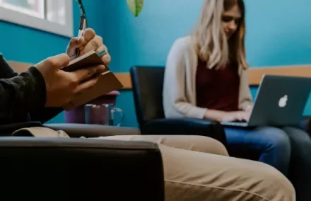Person taking notes while another works on a laptop in a waiting room.