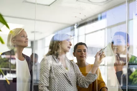 Business meeting with four women discussing in front of a glass board.