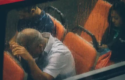 Three people sitting on a bus in the rain, viewed through a wet window.