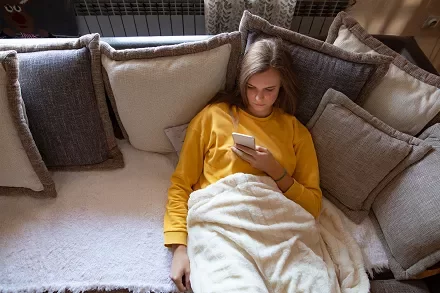 Person in yellow shirt lying on a sofa with pillows, looking at a phone.