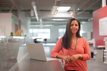 Woman in orange sweater standing by a laptop in a modern office.