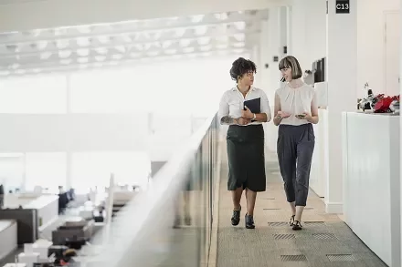 Two women walking and talking in a modern office hallway.