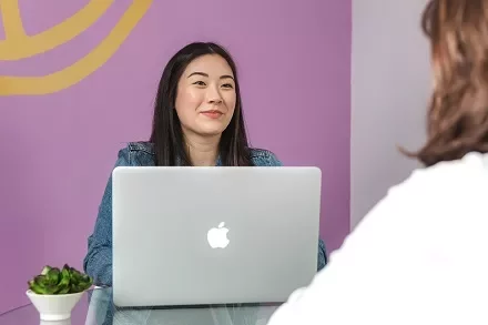 Woman sitting at a table using a laptop, smiling across from another person.