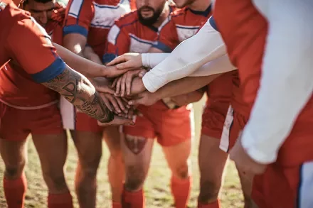 Rugby team huddles with hands together before a match, wearing red uniforms.
