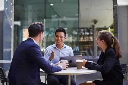 Three people having a meeting at an outdoor cafÃ© table, smiling with coffee cups and a laptop.