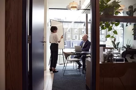 A woman stands at a whiteboard presenting to a seated man in an office with large windows.