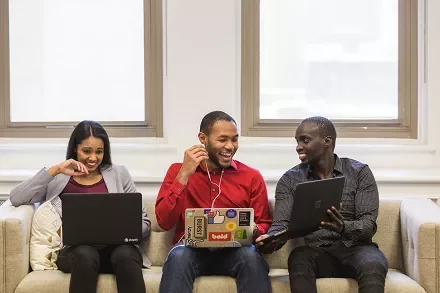 Three people sitting on a couch using laptops, smiling and talking to each other.